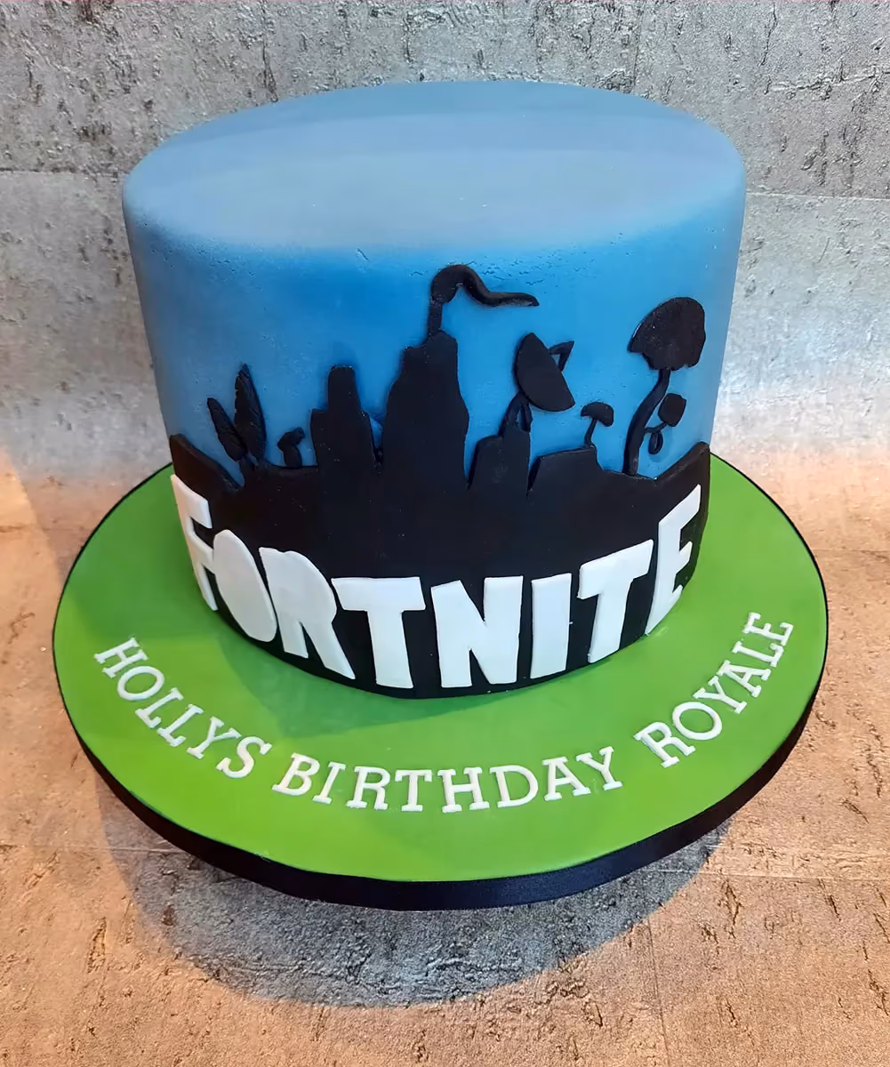 A round, single-tier Fortnite-themed birthday cake. The cake has a light blue top and a black silhouette design of Fortnite landscape elements around its side, with the word 'FORTNITE' in bold white letters. The cake sits on a bright green board, which has 'HOLLYS BIRTHDAY ROYALE' written in white fondant letters around its edge.