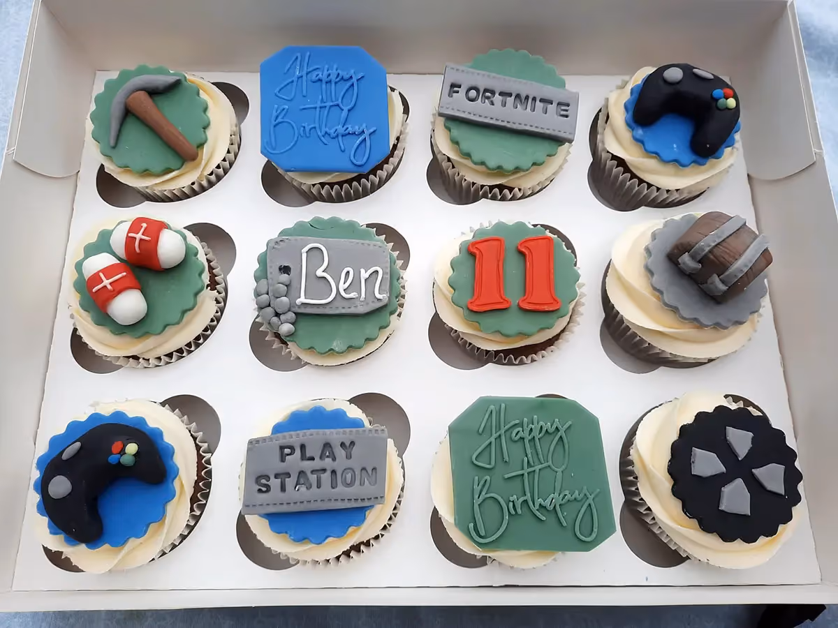 A white box containing twelve cupcakes, each with white frosting and a themed fondant topper. Decorations include gaming controllers, a pickaxe, medical kits, a treasure chest, text reading 'FORTNITE', 'PLAY STATION', 'Happy Birthday', 'Ben', and the number '11', indicating a gaming and Fortnite-themed birthday celebration.