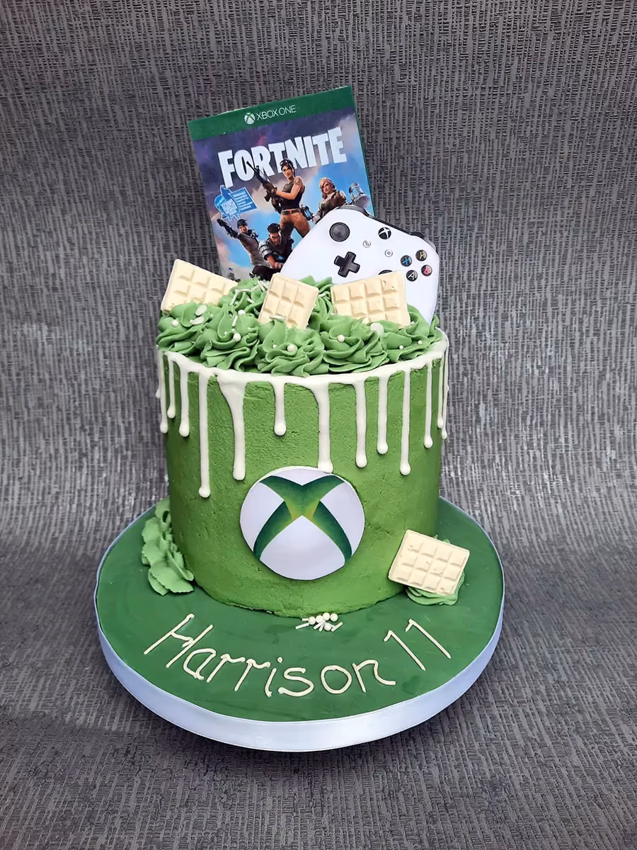 A vibrant green birthday cake with white drip icing, featuring an Xbox logo on the side and topped with a Fortnite game box, an Xbox controller, and white chocolate squares. The green cake board is decorated with white icing spelling 'Harrison 11'.