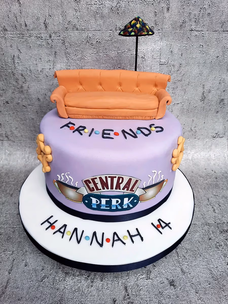 A two-tier Friends-themed birthday cake with a light purple base, topped with an orange couch and a miniature stained-glass lamp. The cake features the "FRIENDS" logo with colored dots, the "CENTRAL PERK" logo, and is personalized with "HANNAH 14" on the white cake board.