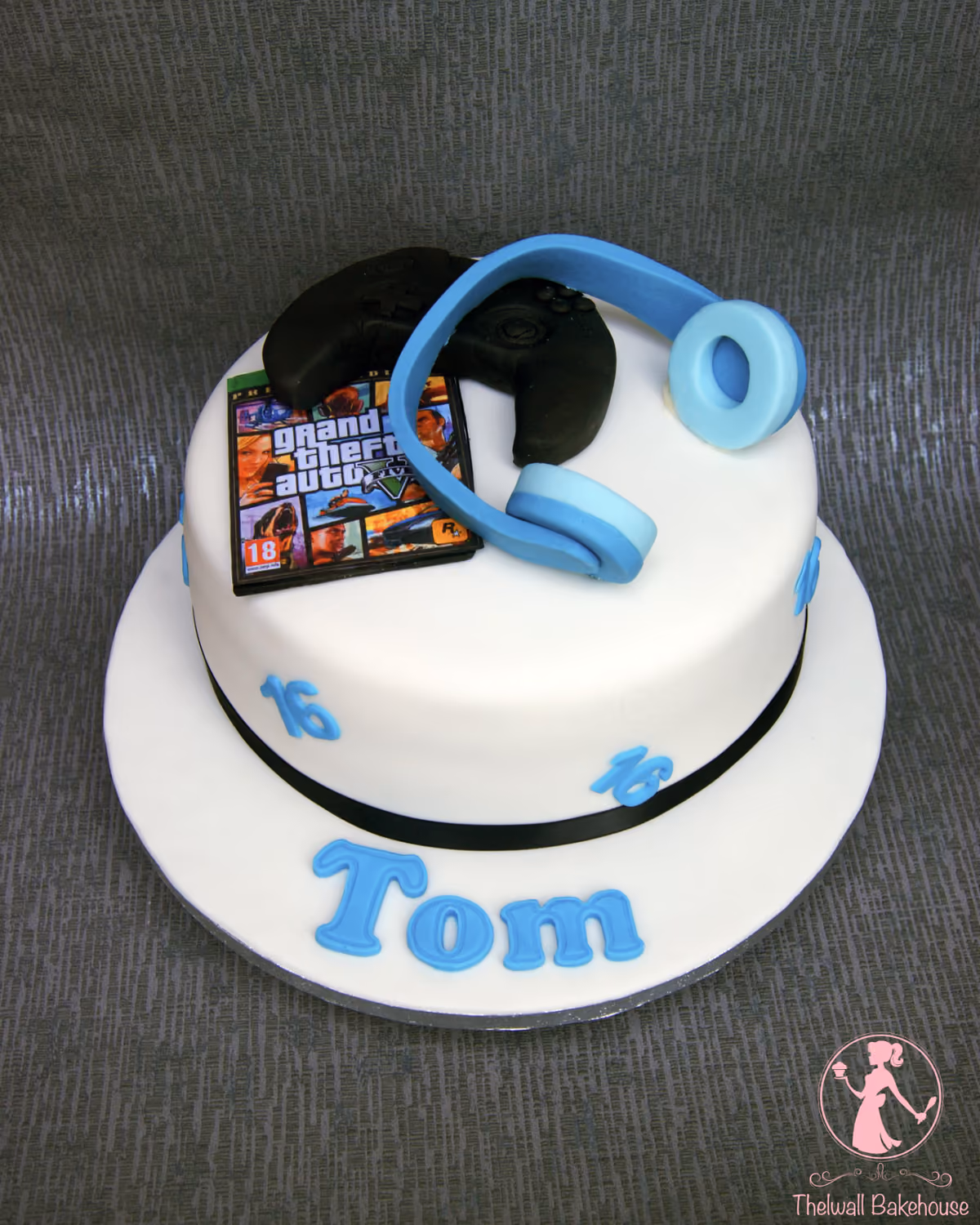 A white round birthday cake decorated with a Grand Theft Auto V game cover, black game controllers, blue headphones, blue number 16s, and the name Tom in blue letters, with a black ribbon around the base.