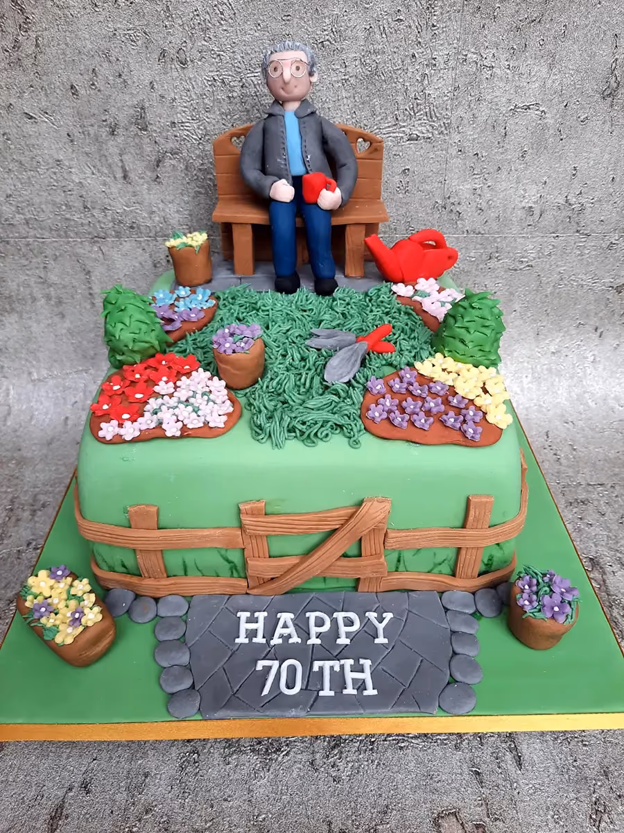 A multi-tier green birthday cake decorated with a detailed garden theme, featuring a figure of an older man with glasses sitting on a wooden bench, surrounded by various potted flowers, gardening tools, and a wooden fence, with a paved path at the base reading 'HAPPY 70TH'.