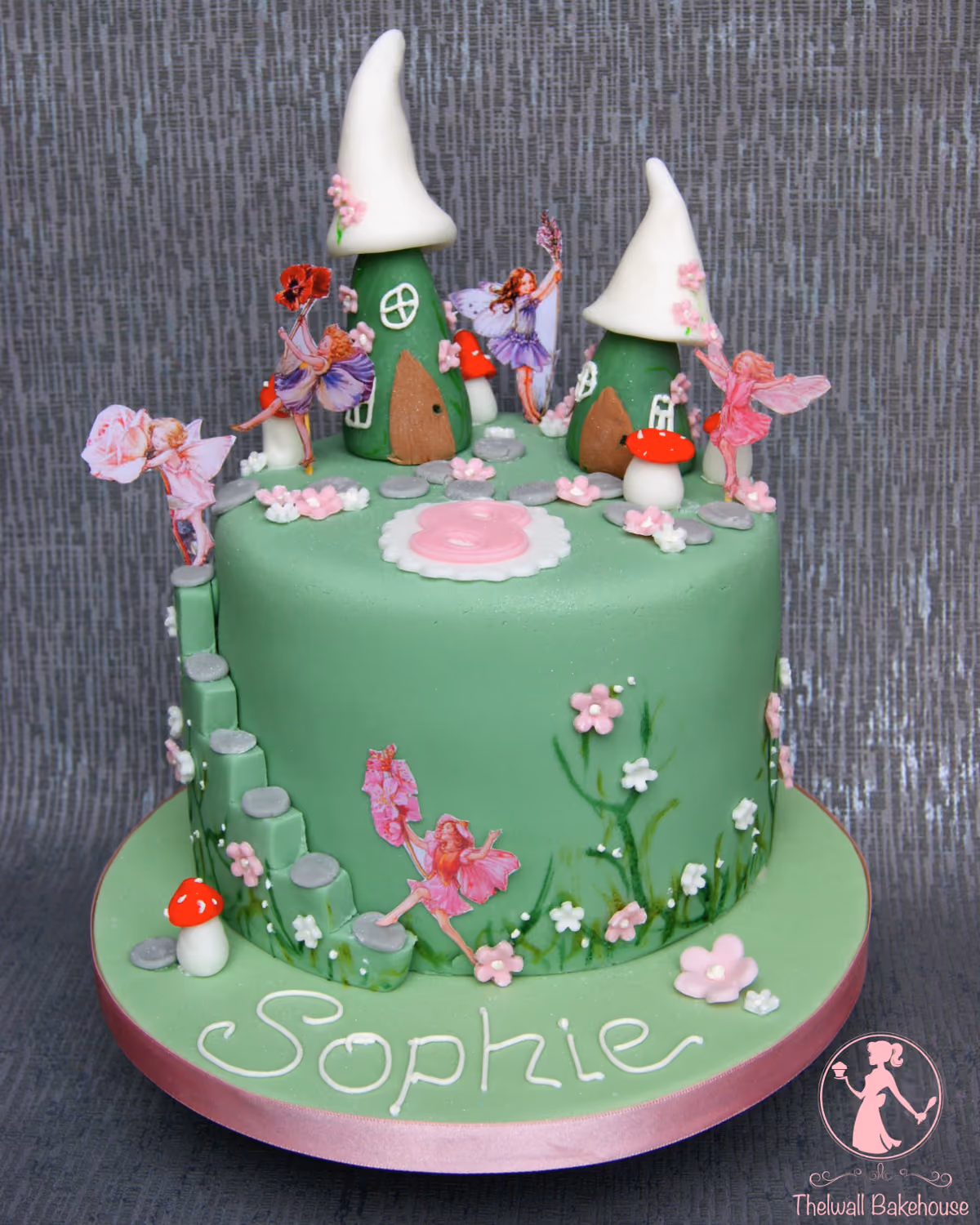 A round, single-tier green fairy garden cake featuring a raised platform with two mushroom houses, decorated with paper cutout fairies, sugar mushrooms, flowers, and a side staircase, with the name 'Sophie' written in white on the base.