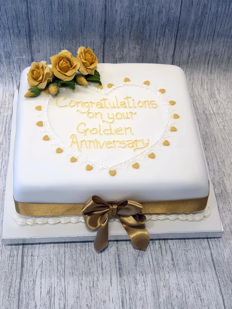 A square golden anniversary cake with white fondant, decorated with three yellow roses, gold hearts, and a gold ribbon with a bow. The cake's top features the message 'Congratulations on your Golden Anniversary' in gold icing, framed by a white lace heart.