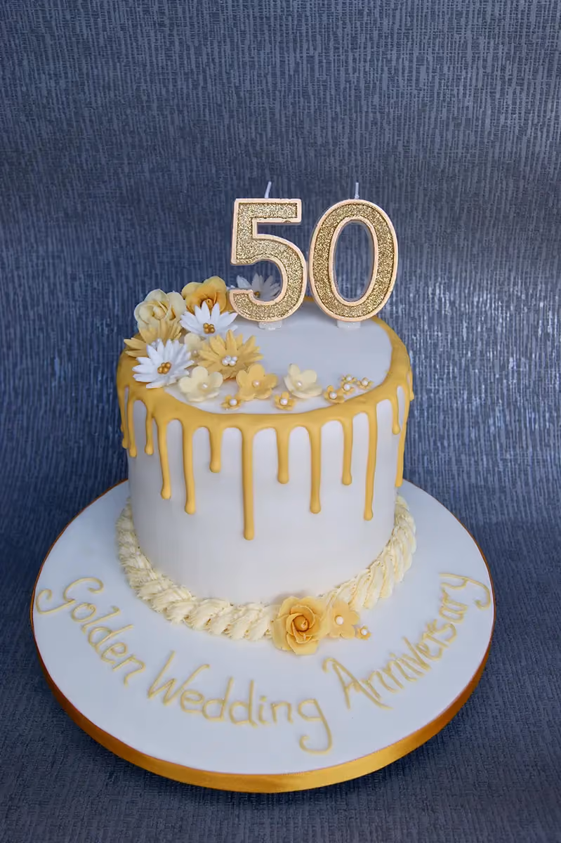 A white single-tier cake with gold drip icing, topped with glittery gold number 50 candles and decorated with yellow and white sugar flowers. The cake board has "Golden Wedding Anniversary" written in gold.