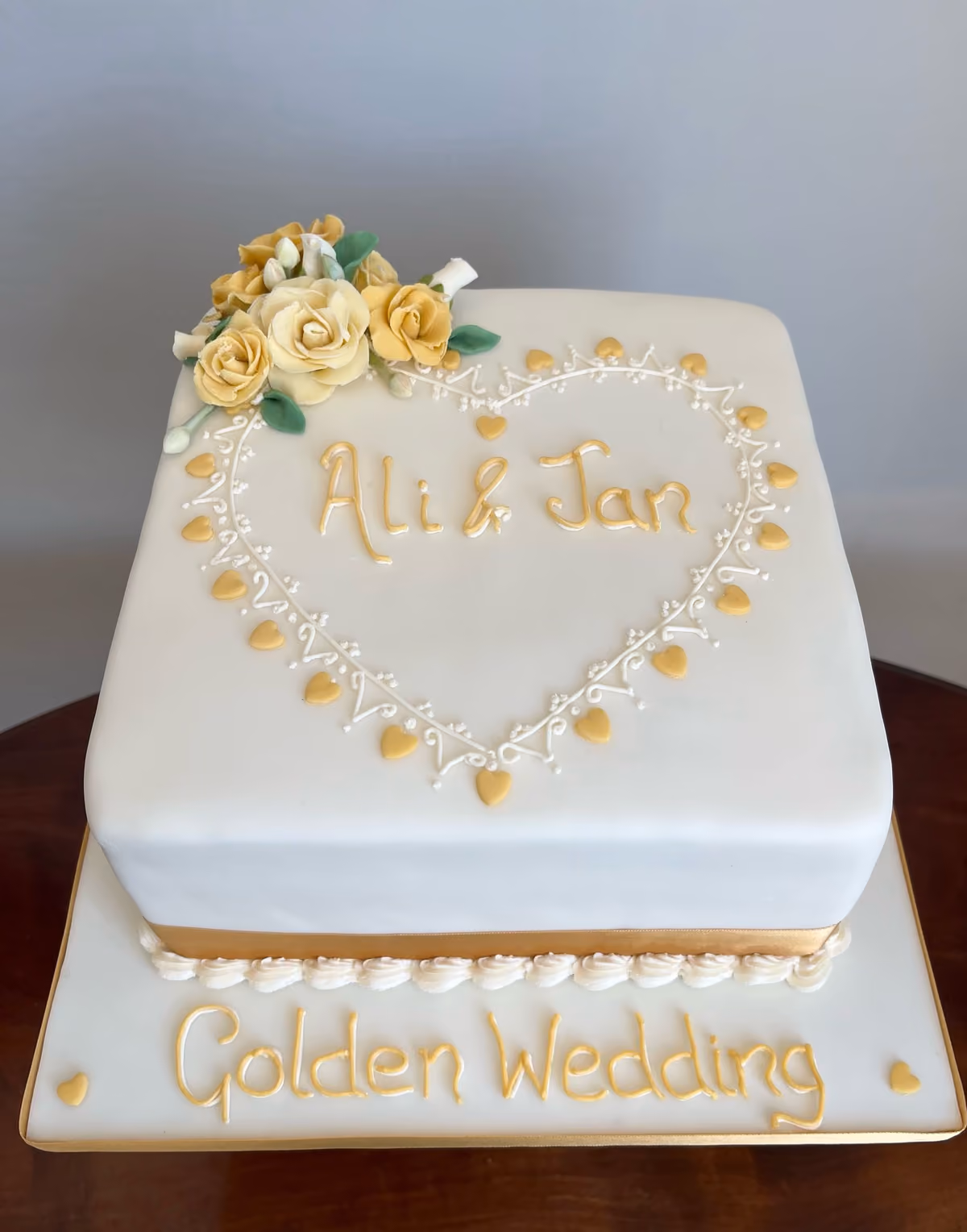 A square white fondant Golden Wedding Anniversary cake, decorated with yellow sugar roses and green leaves on top. A heart outline made of white piped icing and golden hearts encloses the names 'Ali & Jan' in gold script. The cake board features 'Golden Wedding' in gold script with small golden hearts.