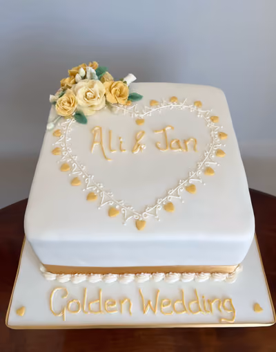 A square white fondant Golden Wedding Anniversary cake, decorated with yellow sugar roses and green leaves on top. A heart outline made of white piped icing and golden hearts encloses the names 'Ali & Jan' in gold script. The cake board features 'Golden Wedding' in gold script with small golden hearts.