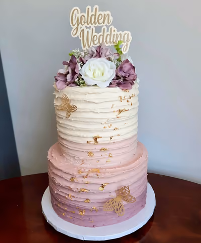 Two-tier Golden Wedding cake with white and ombre pink to purple textured frosting, decorated with gold leaf flakes, gold butterflies, and a floral arrangement with a 'Golden Wedding' topper.