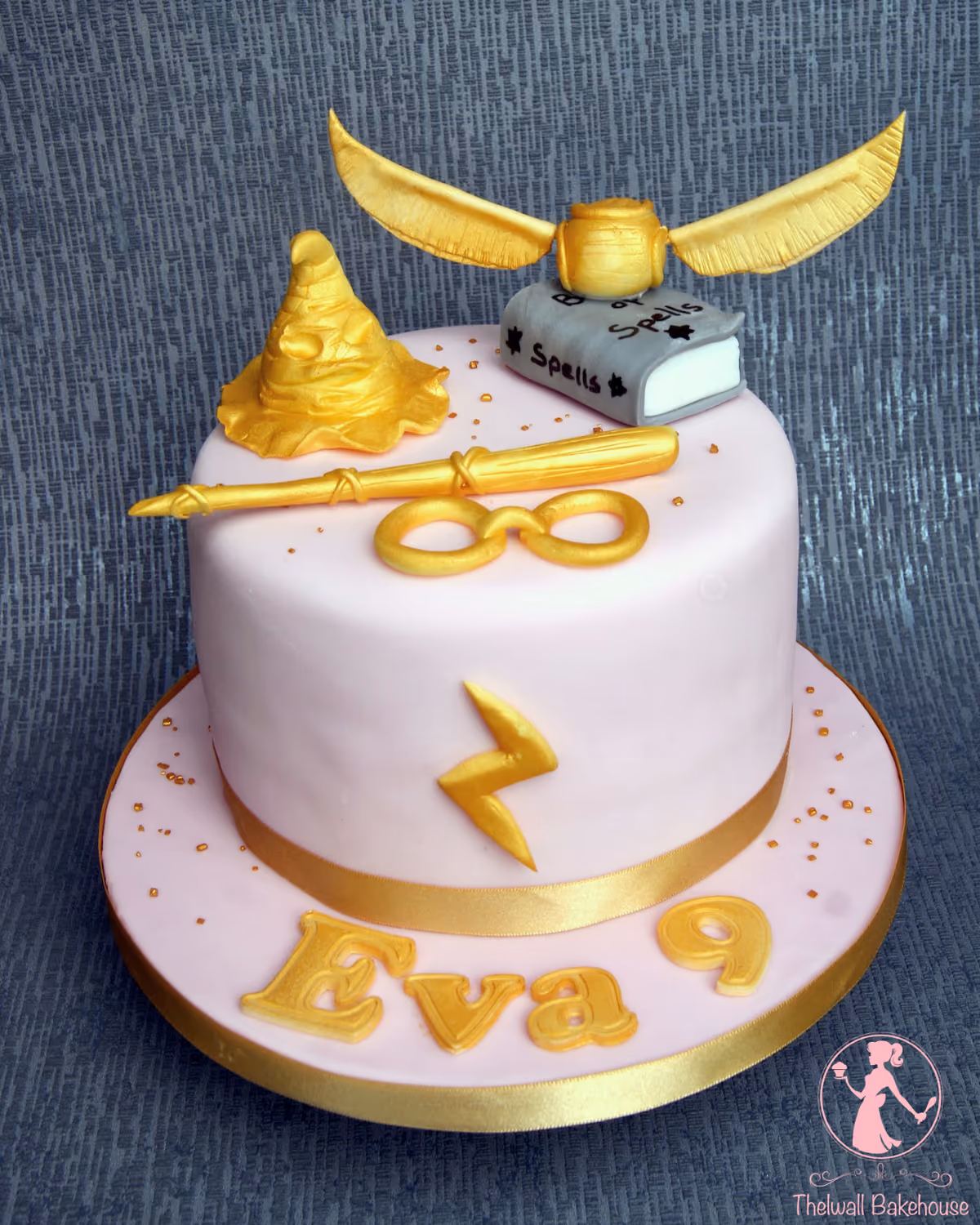 A light pink, single-tier Harry Potter themed birthday cake for Eva's 9th birthday, decorated with gold elements including a Golden Snitch, a sorting hat, a wand, glasses, a lightning bolt, and a grey 'Book of Spells' on top, with 'Eva' and '9' written in gold on the cake board.