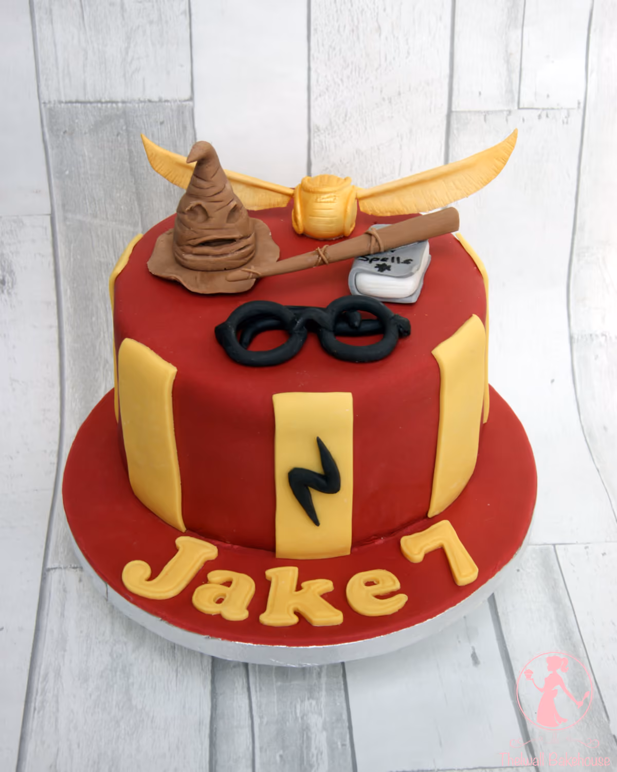 A single-tier Harry Potter themed birthday cake with red and yellow fondant, decorated with edible toppers including the Sorting Hat, a Golden Snitch, a magic wand, a 'Spells' book, Harry Potter's round glasses, and a lightning bolt. The cake board features the name 'Jake' and the number '7' in yellow fondant.