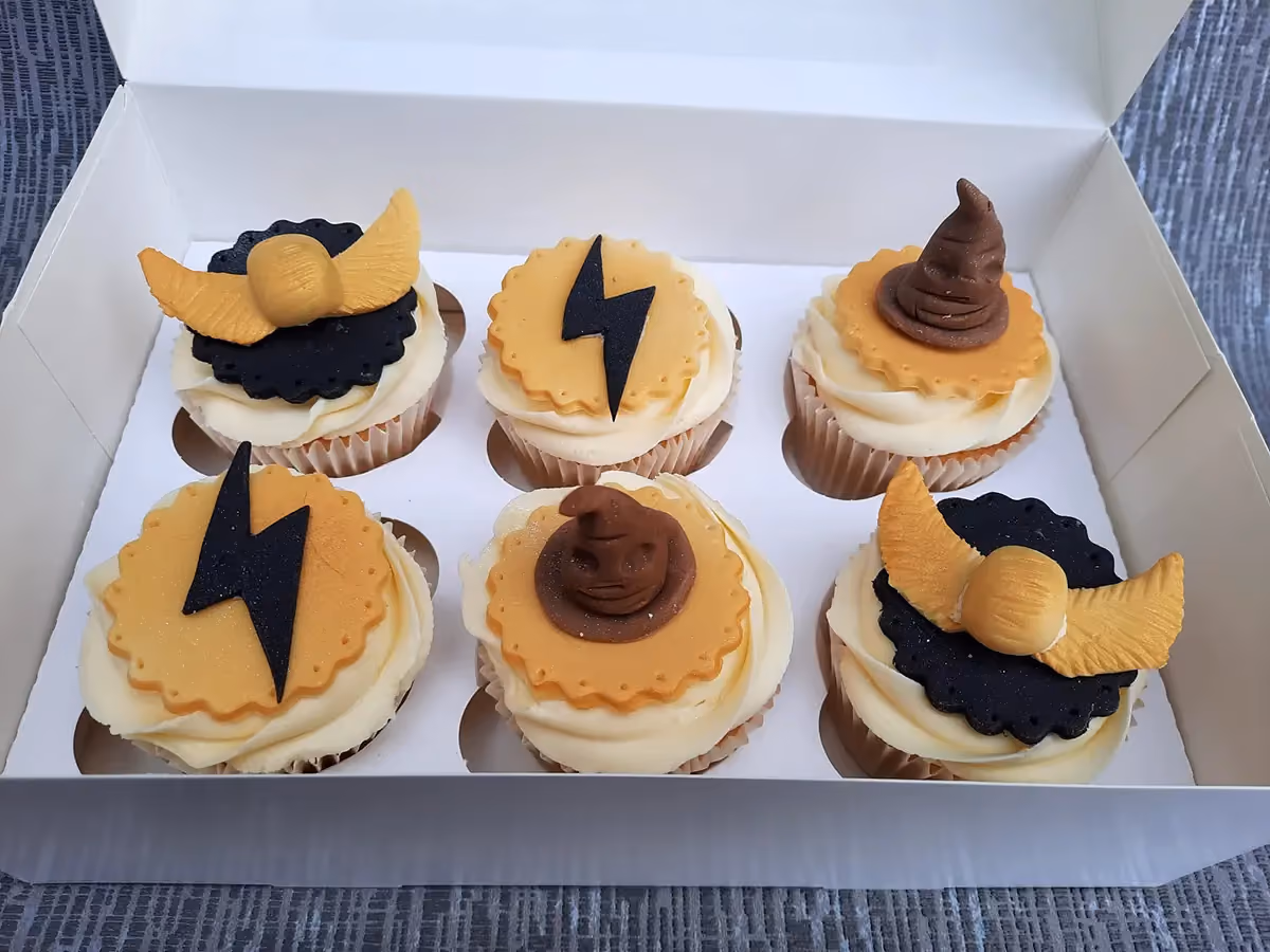 Six Harry Potter themed cupcakes in a white box, featuring golden snitch, lightning bolt, and sorting hat decorations on vanilla frosting.