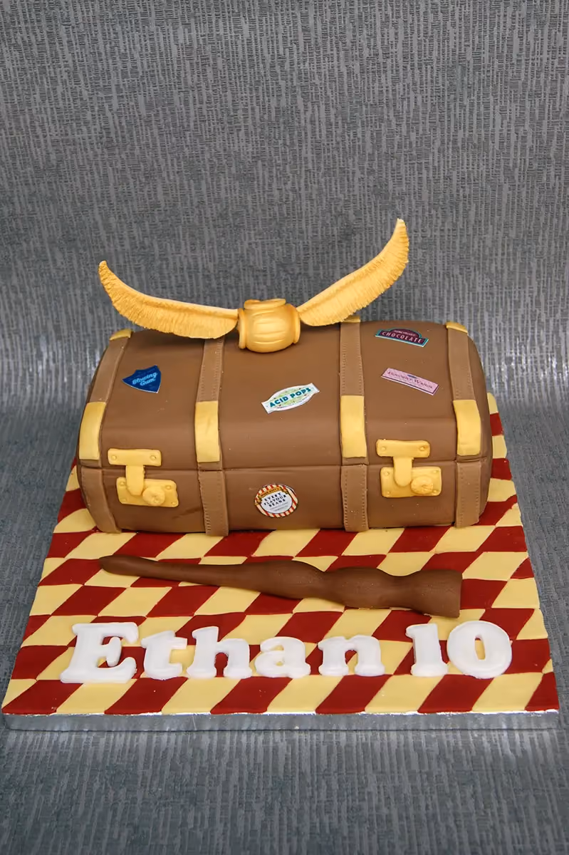 A Harry Potter themed cake shaped like a brown suitcase, decorated with a golden snitch on top, a brown wand, and the name 'Ethan 10' in white letters on a red and yellow checkered base.