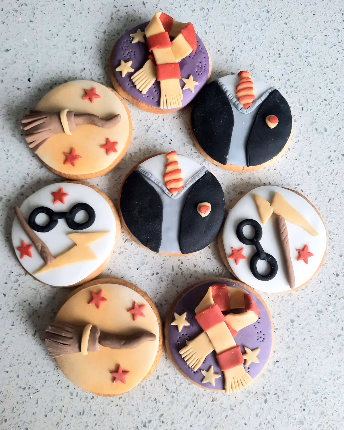 Seven round cookies decorated with Harry Potter themed fondant designs, including a Gryffindor scarf, broomstick, Harry Potter's glasses and lightning scar, wizard robes with a tie, and Quidditch elements.