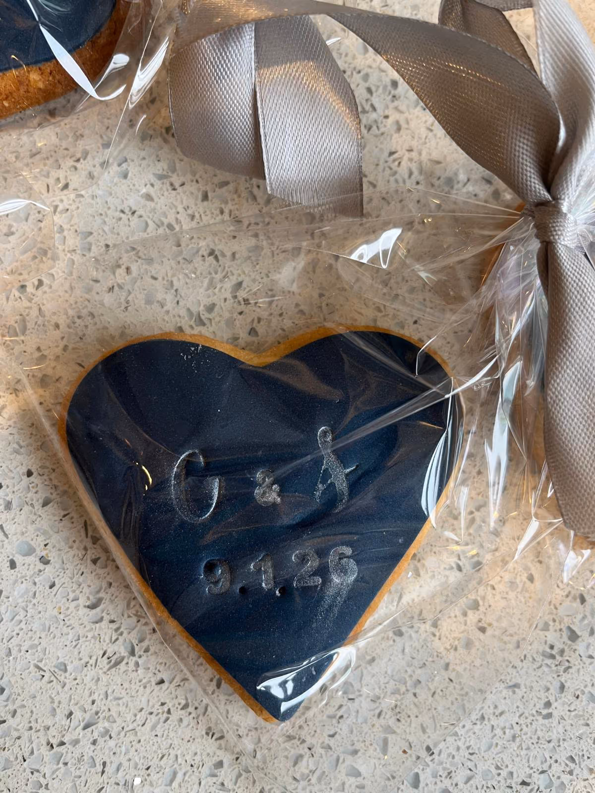 A heart-shaped biscuit with navy blue icing and silver text 'G & A 9.1.26', individually wrapped in clear plastic and tied with a grey ribbon, resting on a speckled countertop.