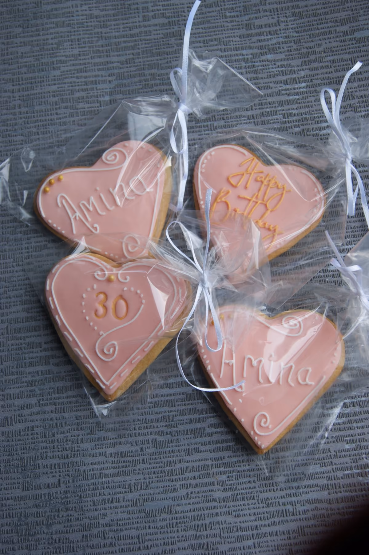 Four pink heart-shaped iced cookies, individually wrapped with white ribbons. Two cookies are personalized with 'Amina', one says 'Happy Birthday', and one features the number '30'.