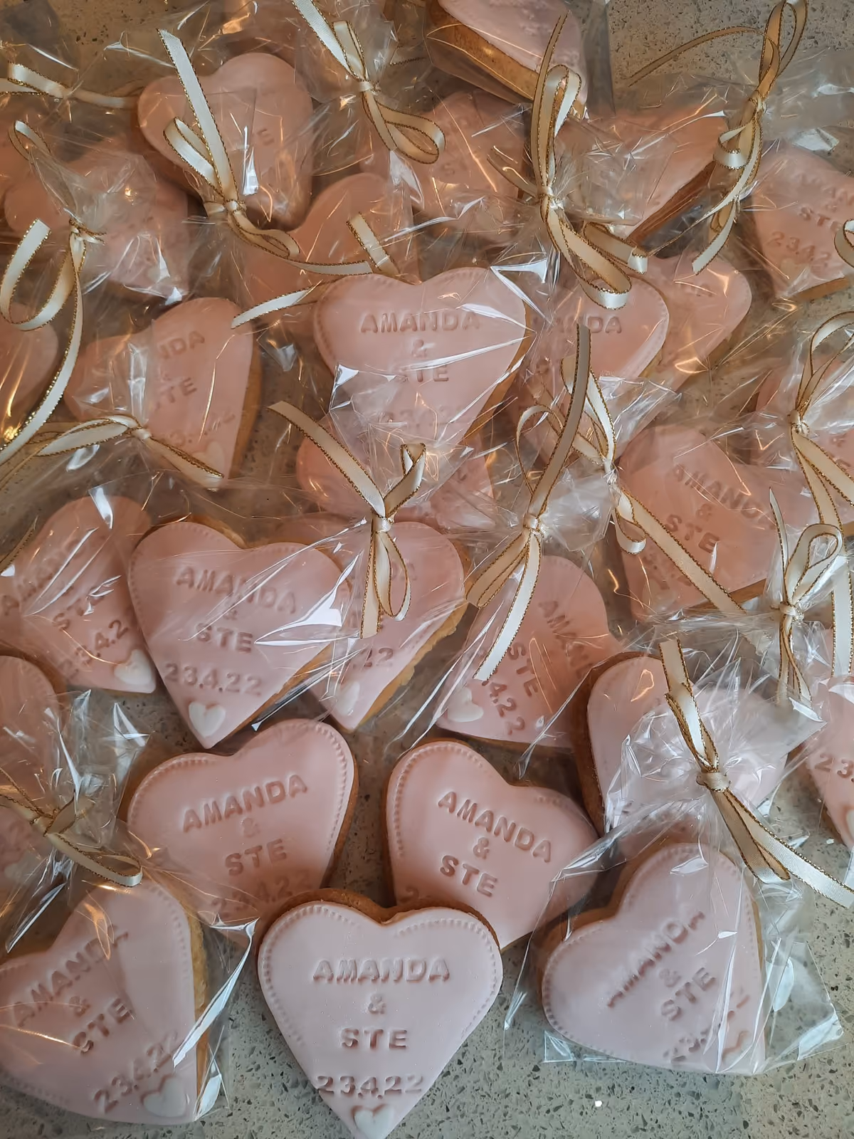 A close-up overhead view of numerous heart-shaped cookies, each individually wrapped in clear plastic and tied with a white and gold ribbon. The light pink iced cookies are personalized with 'AMANDA & STE' and '23.4.22', along with a small white heart at the bottom.