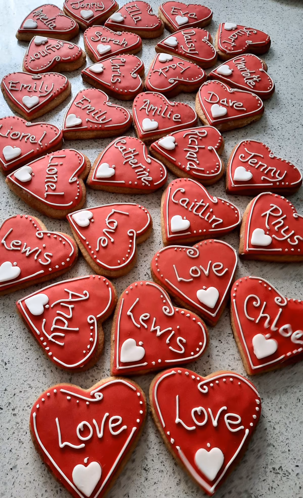 A collection of heart-shaped cookies with vibrant red icing, each intricately decorated with a personalized name or the word "Love" in white icing, and a small white heart accent.
