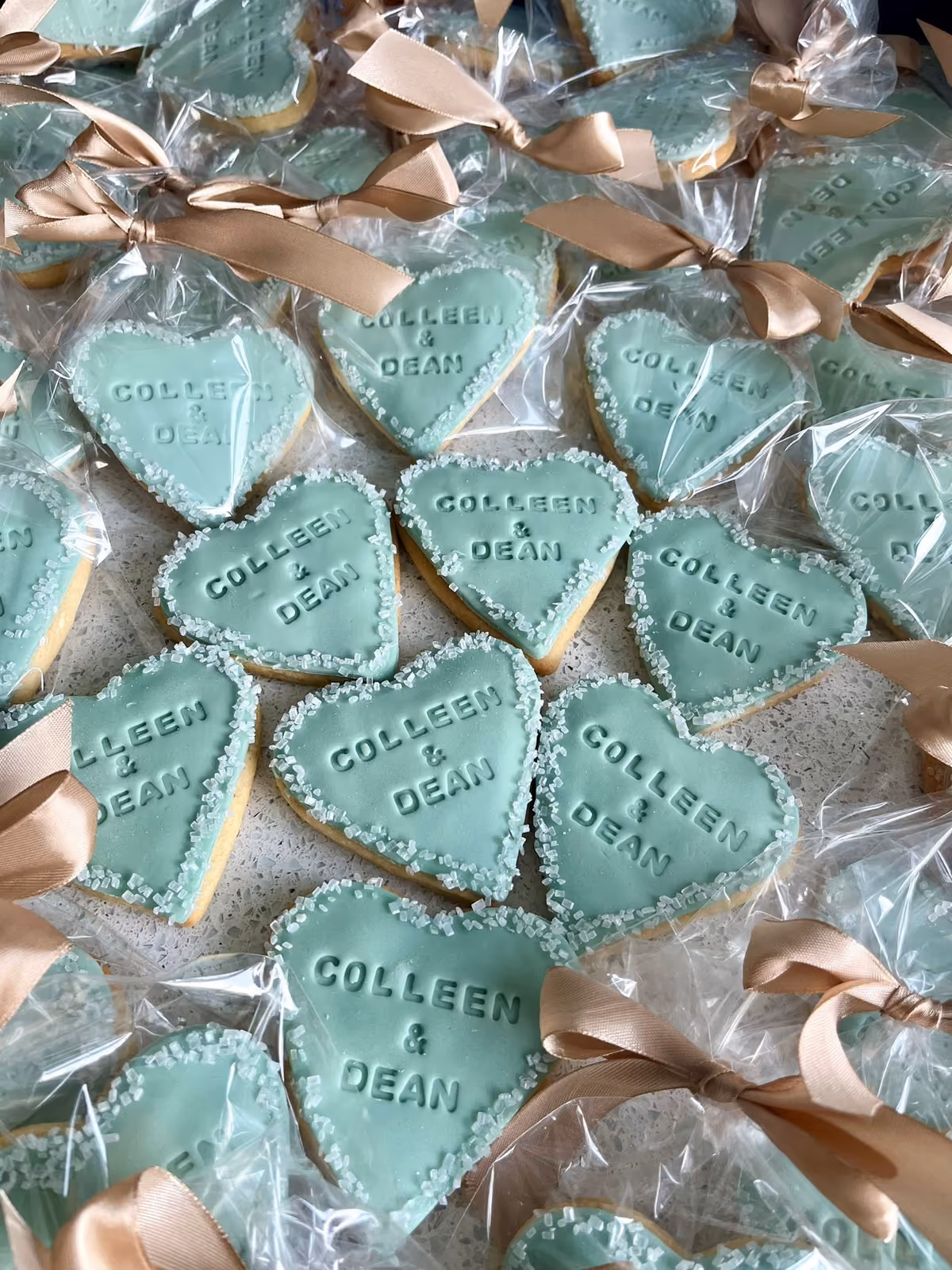 A close-up view of numerous teal heart-shaped cookies, each embossed with the names 'COLLEEN & DEAN' and edged with white sugar sprinkles, individually wrapped in clear plastic bags tied with a beige ribbon.