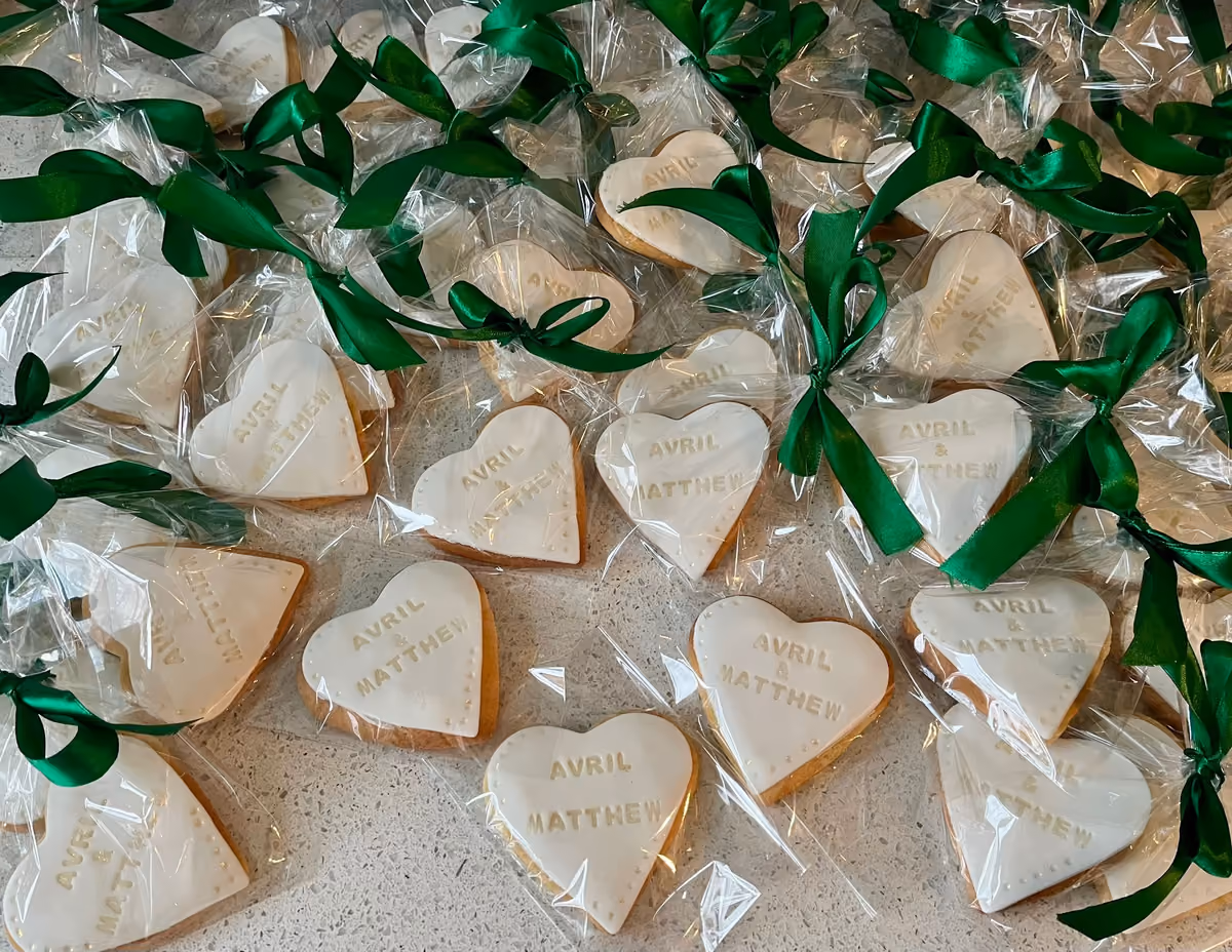 A flat lay of many heart-shaped white-iced biscuits, each individually wrapped in clear plastic and tied with a green satin ribbon. The biscuits are personalised with the names 'Avril & Matthew' in gold lettering, and some have small decorative dots around the edges.