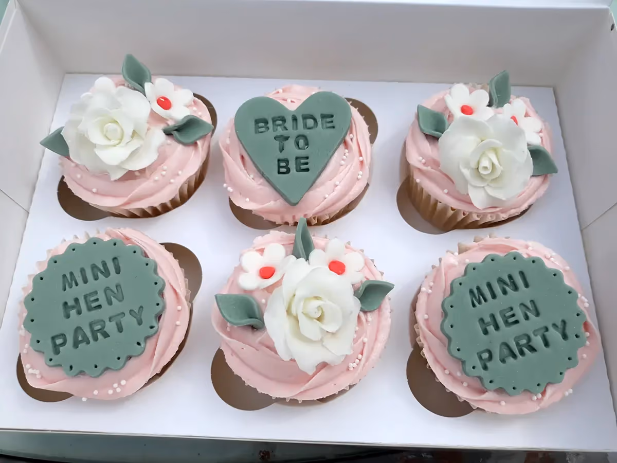 Six pink frosted cupcakes in a white box, three decorated with white roses, white flowers, and green leaves. One cupcake has a green heart-shaped 'BRIDE TO BE' topper, and two have round green 'MINI HEN PARTY' toppers, all with white sprinkles.