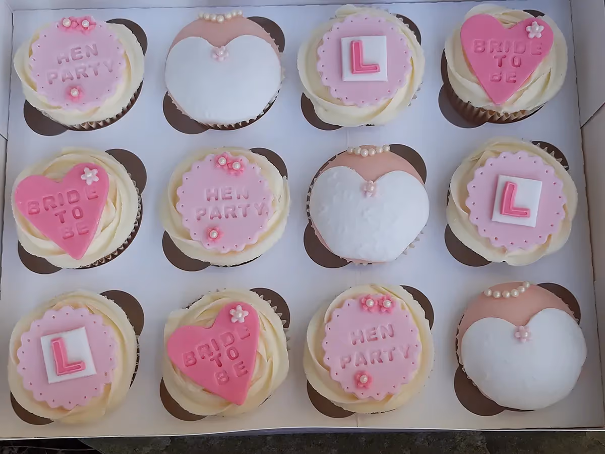 A dozen pink and white hen party themed cupcakes displayed in a white box. Designs include pink toppers with 'HEN PARTY' or 'BRIDE TO BE' text, a pink letter 'L' on a white square, and cupcakes decorated to resemble a white and pink dress with pearl accents.