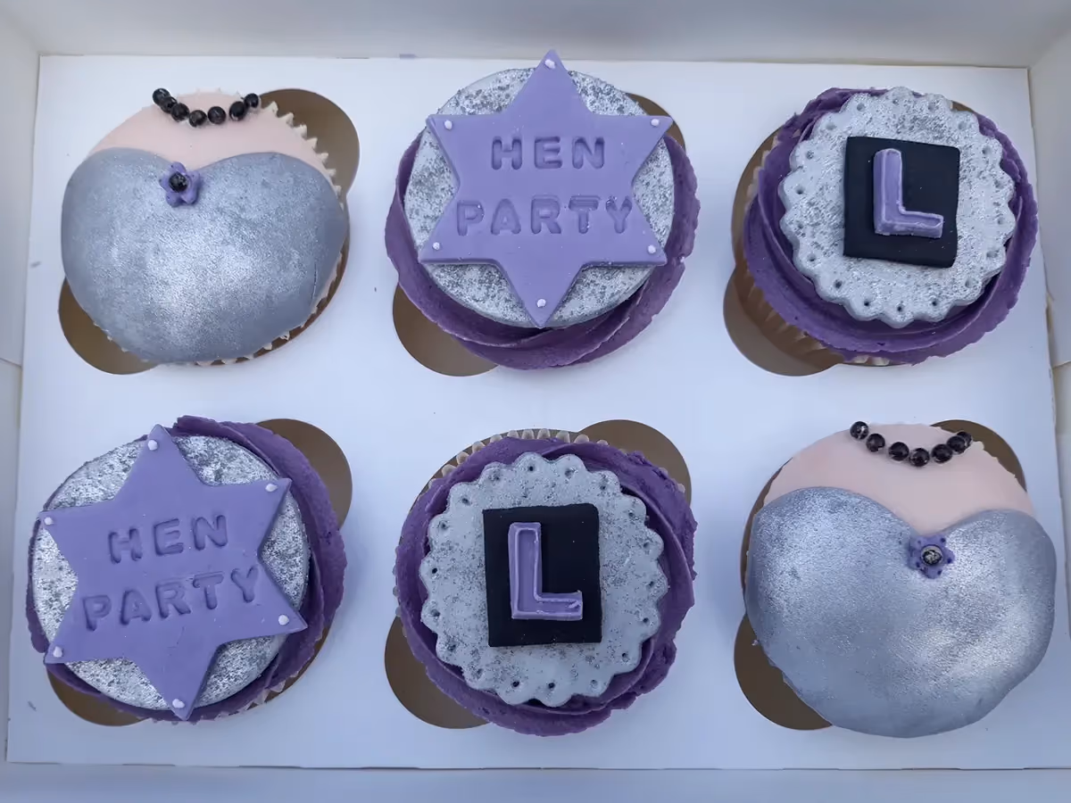 Six hen party themed cupcakes in a white box. The cupcakes feature three distinct designs: two are shaped like a silver and pink dress bodice with a black bead necklace, two have a purple star with 'HEN PARTY' text on a silver base, and two have a purple letter 'L' on a black square on a silver ruffled base. All are set on purple frosting.