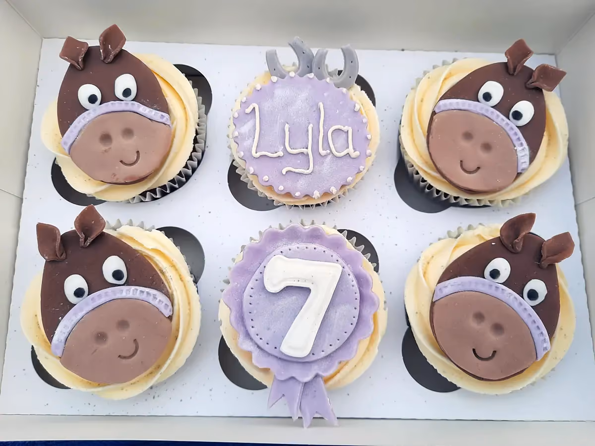 Six horse-themed cupcakes in a white box, featuring four cupcakes with brown horse faces, one purple cupcake with 'Lyla' written in white, and one purple rosette cupcake with a white number 7.