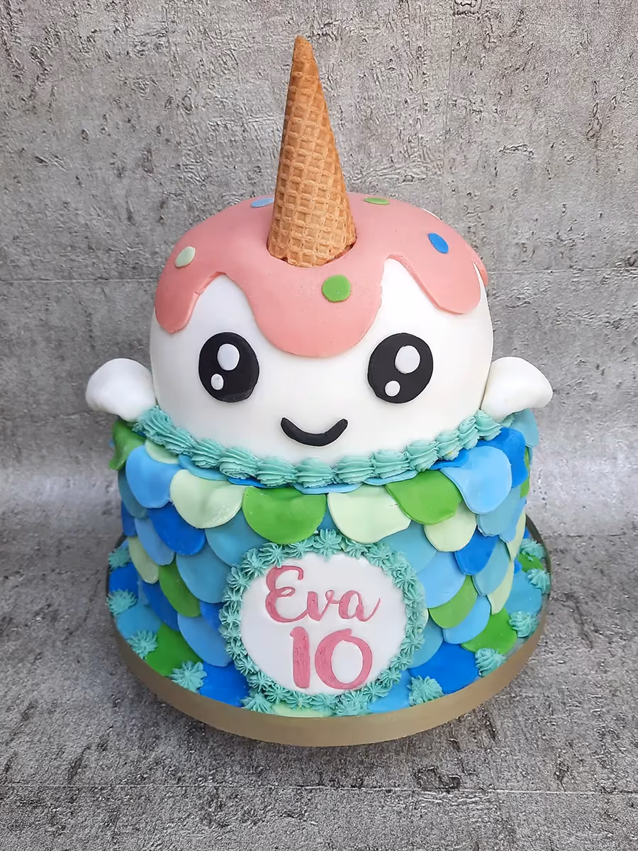 A two-tier birthday cake featuring a cute white character with a pink ice cream drip and an ice cream cone horn on the top tier, and a bottom tier decorated with blue and green mermaid-like scales, with a white plaque displaying 'Eva 10' in pink.