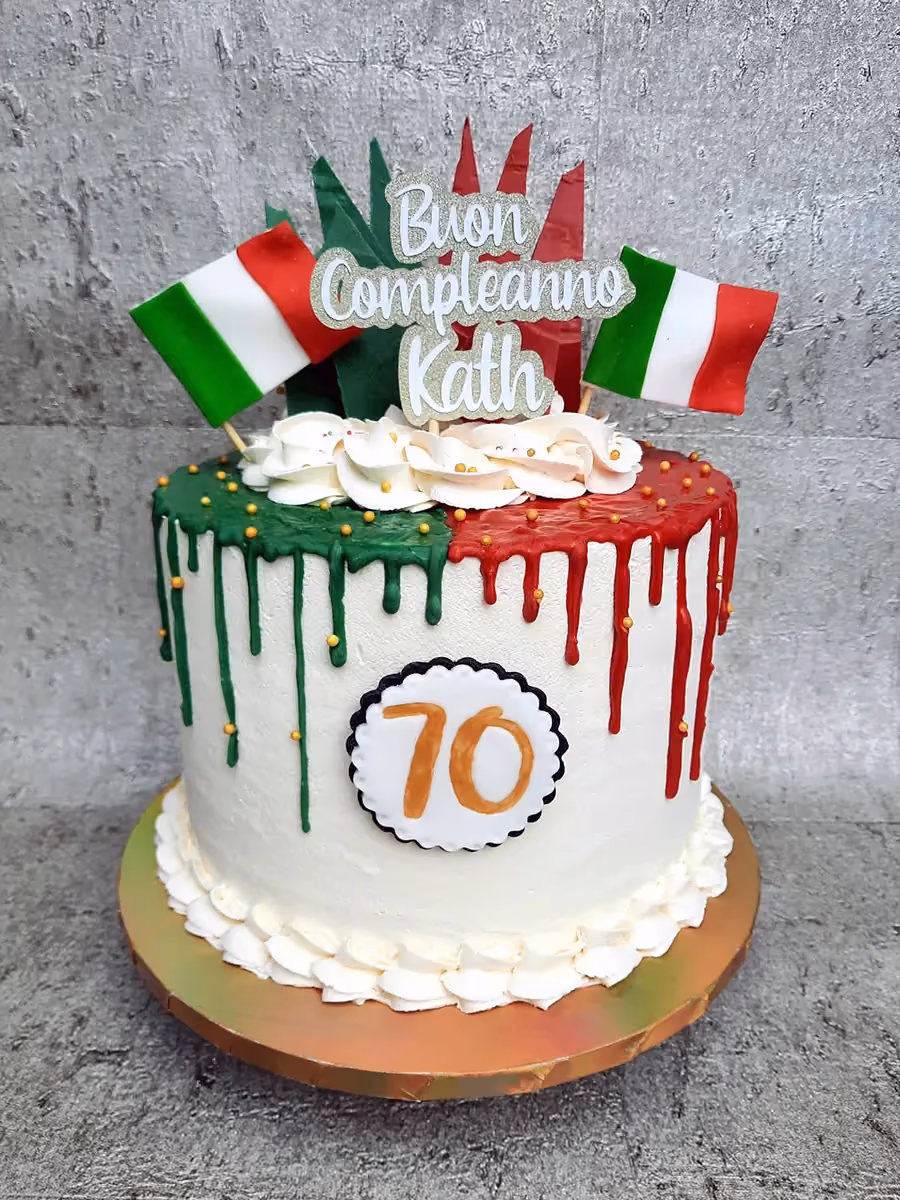 A white birthday cake decorated with Italian flag colors, featuring green and red drip icing on top, two miniature Italian flag toppers, a 'Buon Compleanno Kath' topper, and the number '70' on a white and black scalloped disc on the side.