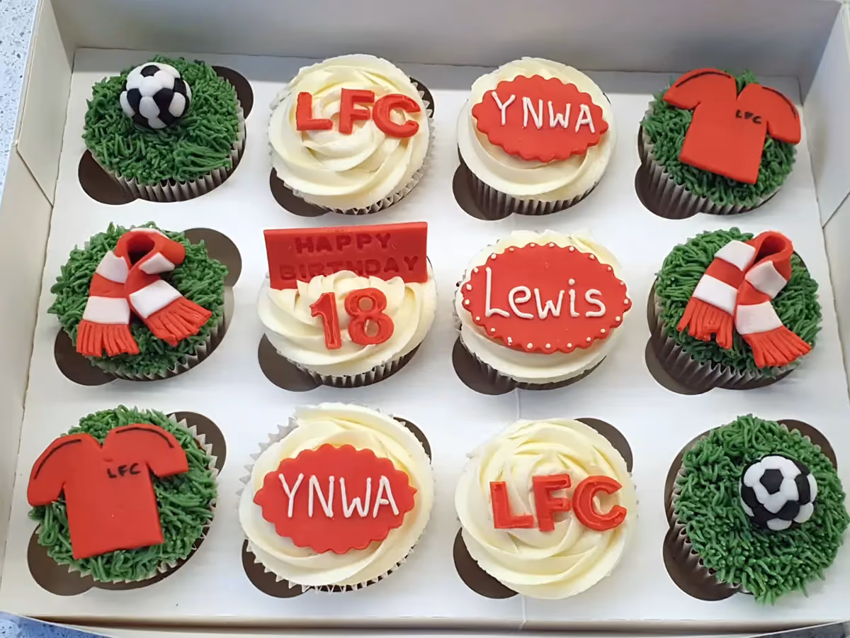 A dozen Liverpool FC themed cupcakes in a white box. Decorations include green grass icing with footballs and red and white scarves, and white icing with red toppers spelling 'LFC', 'YNWA', 'Lewis', and 'Happy Birthday 18'.