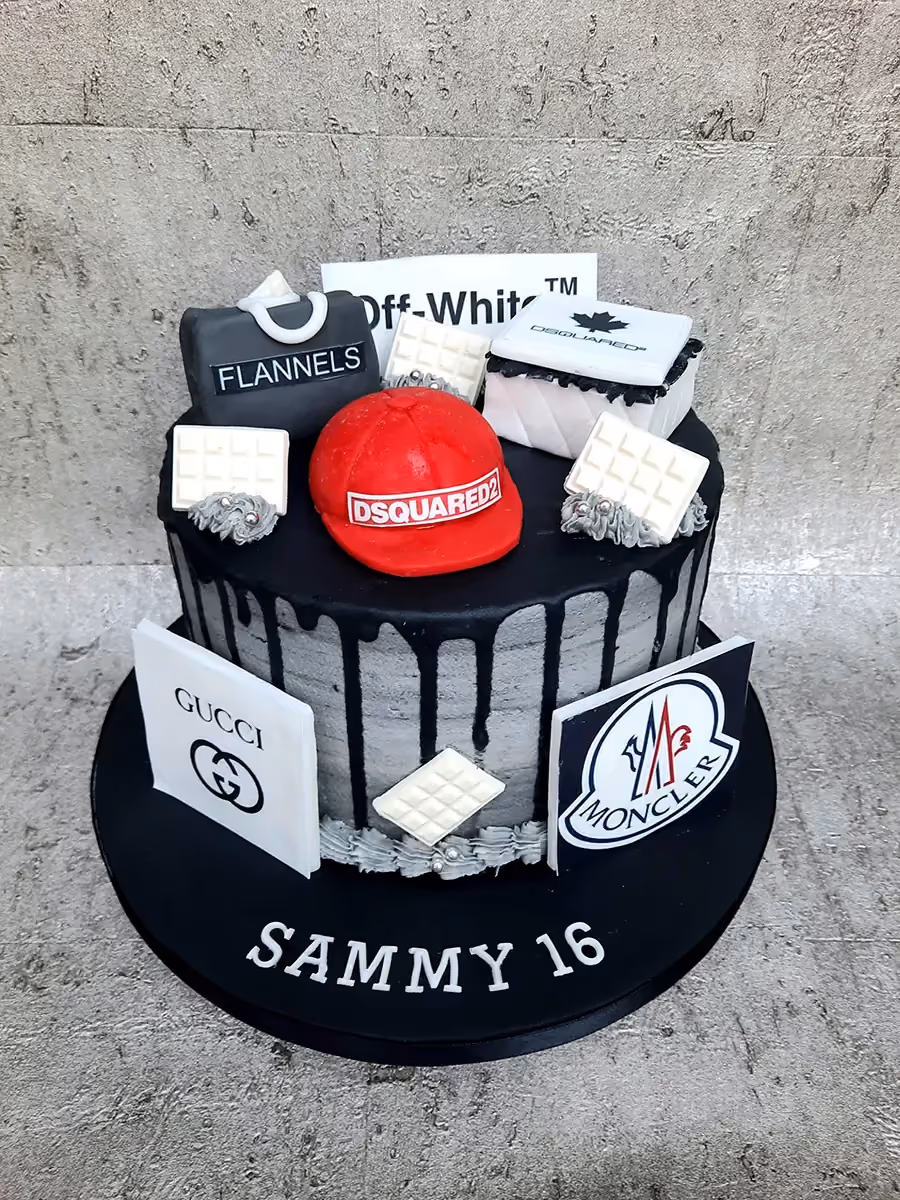 A single-tier birthday cake decorated with luxury fashion brand logos and items, including Off-White, Flannels, Dsquared2, Gucci, and Moncler, featuring black drip icing over grey textured sides, and 'SAMMY 16' written on the black cake board.
