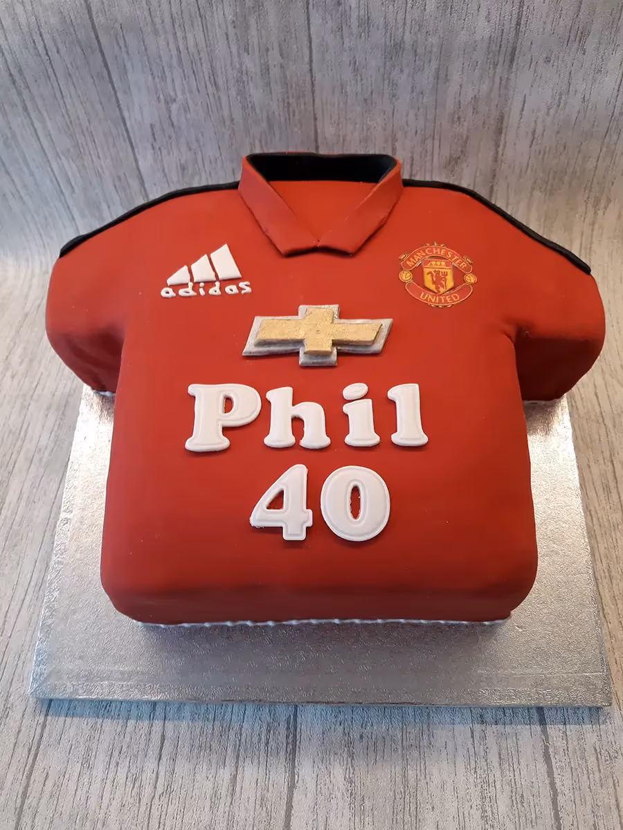 A red football shirt shaped cake, decorated with a Manchester United crest, an Adidas logo, a gold Chevrolet logo, and the name 'Phil' and number '40' in white icing.