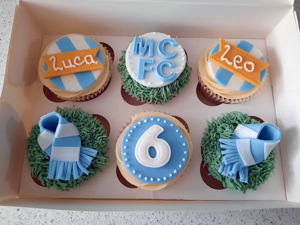 Six Manchester City themed cupcakes in a white box, featuring names 'Luca' and 'Leo', 'MCFC' logo, a number '6', and blue and white striped scarves on green grass-like frosting.
