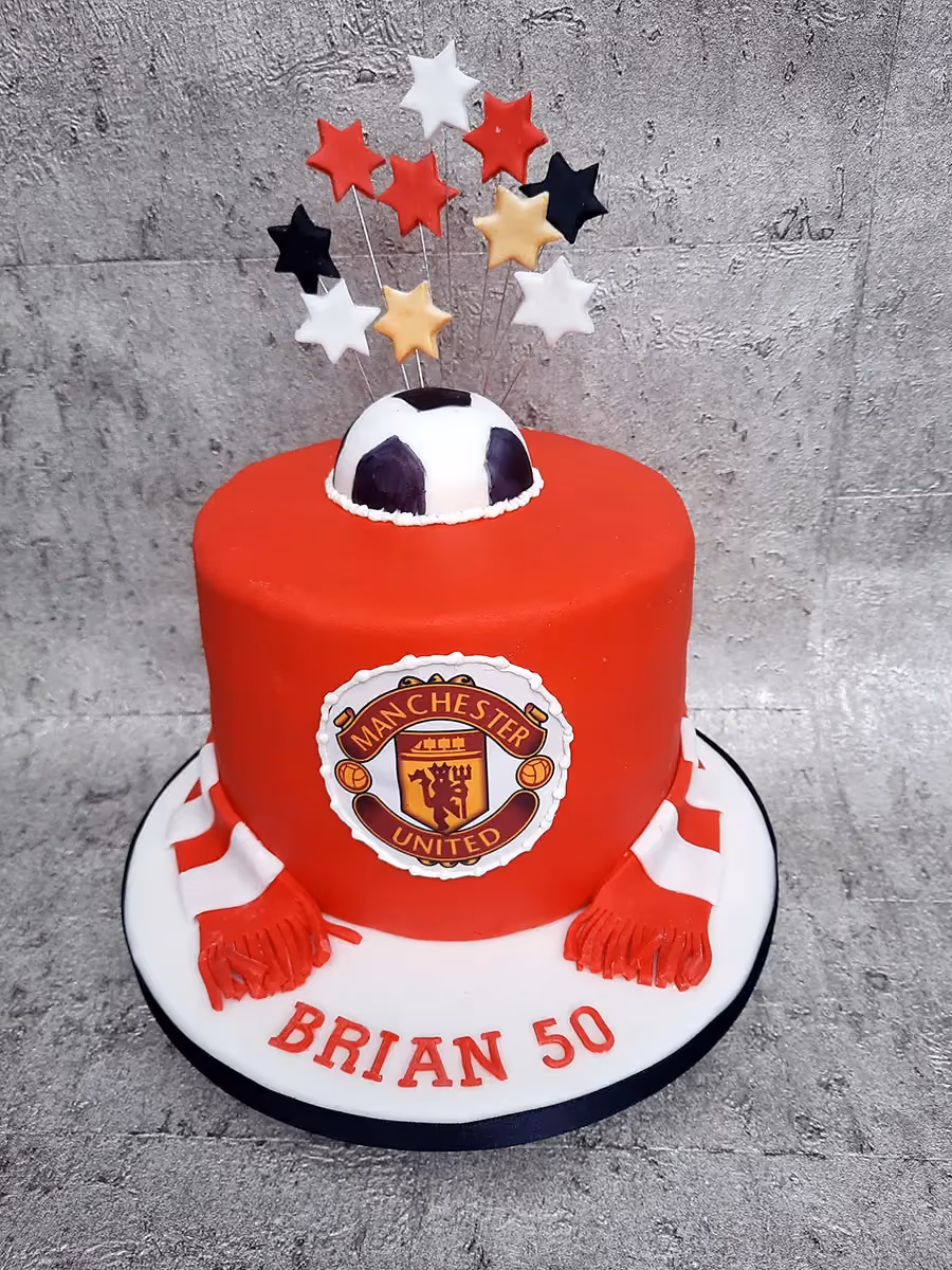 A round red birthday cake decorated with a Manchester United logo on the front, a half-sphere football on top, and an explosion of red, white, black, and gold stars on wires. A red and white striped scarf is draped around the base of the cake, and the words 'BRIAN 50' are written in red on the white cake board.