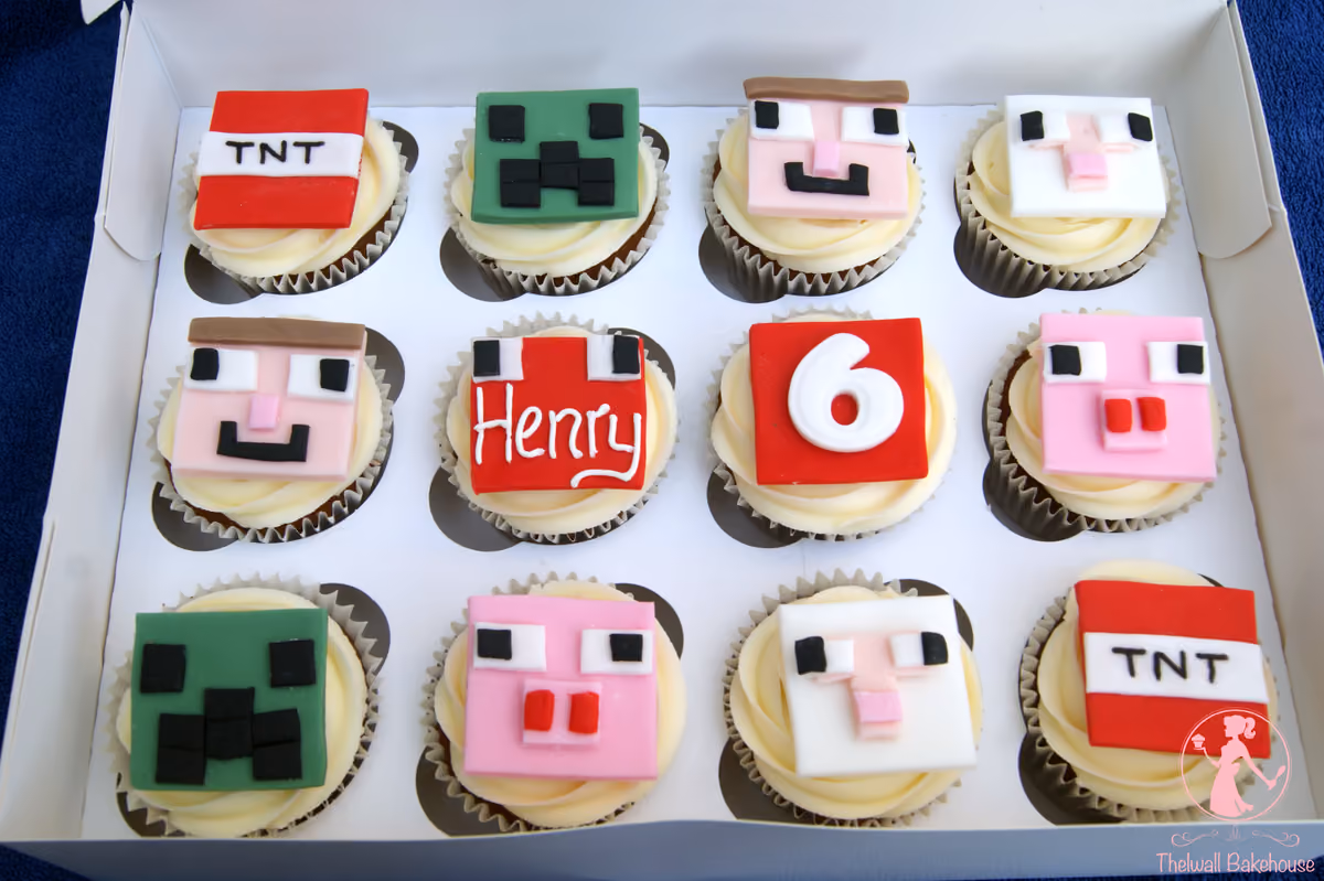A dozen Minecraft-themed cupcakes in a white box, featuring assorted fondant toppers including TNT blocks, Creeper, Steve, Pig, and Sheep faces, with two special cupcakes displaying 'Henry' and the number '6' on red backgrounds.