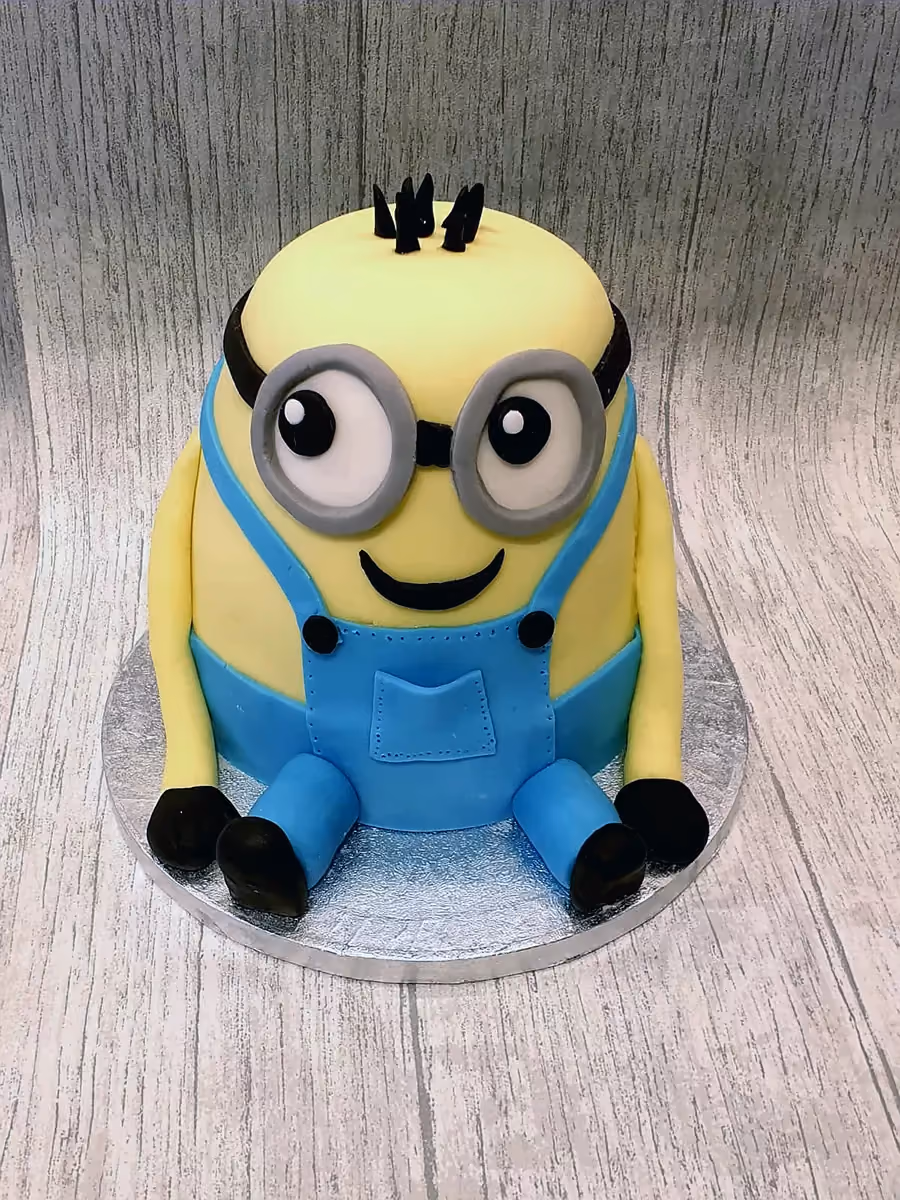 A Minion-shaped cake featuring a yellow body, blue overalls, goggles, and black hair, sitting on a silver cake board.
