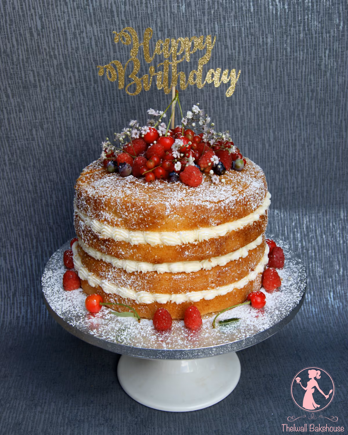 A three-tier naked birthday cake with white cream filling, dusted with powdered sugar, and topped with fresh mixed berries and small white flowers, featuring a gold glitter 'Happy Birthday' topper, displayed on a silver cake stand.