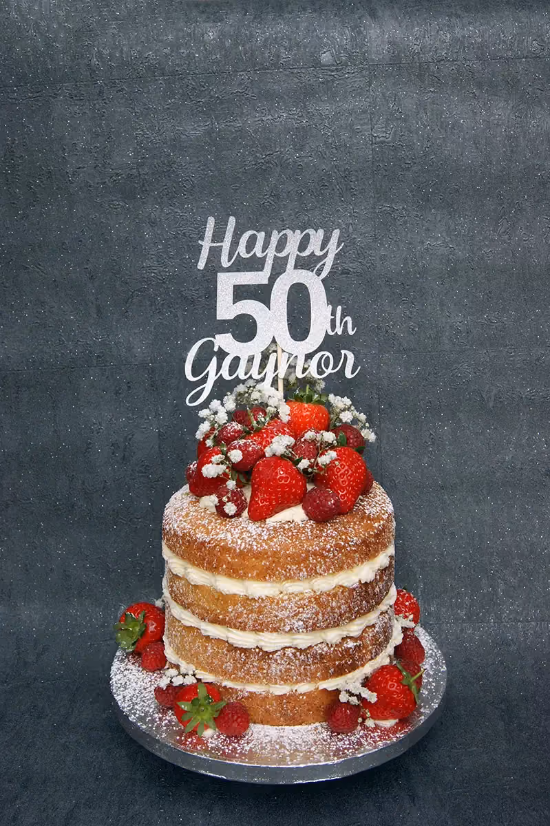A three-tier naked birthday cake with white cream filling, topped with fresh strawberries, raspberries, and small white flowers, dusted with icing sugar, and featuring a silver glitter 'Happy 50th Gaynor' cake topper.