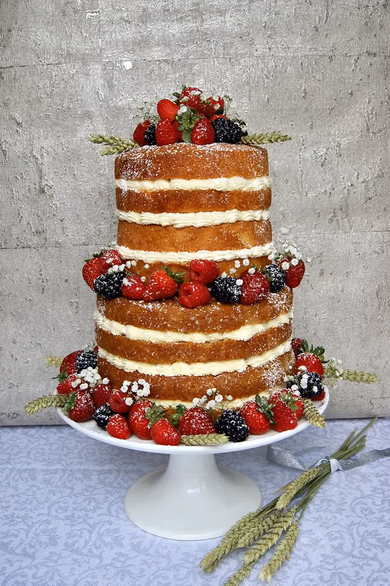A three-tier naked cake featuring visible sponge layers and cream filling, adorned with fresh strawberries, raspberries, blackberries, small white flowers, and wheat stalks, all lightly dusted with powdered sugar, set on a white cake stand.