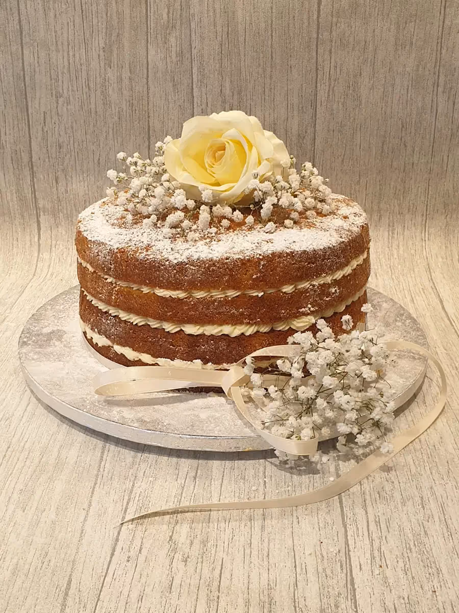 A two-tier naked cake dusted with icing sugar, decorated on top with a yellow rose and baby's breath flowers, and a cream ribbon tied around the base with additional baby's breath.