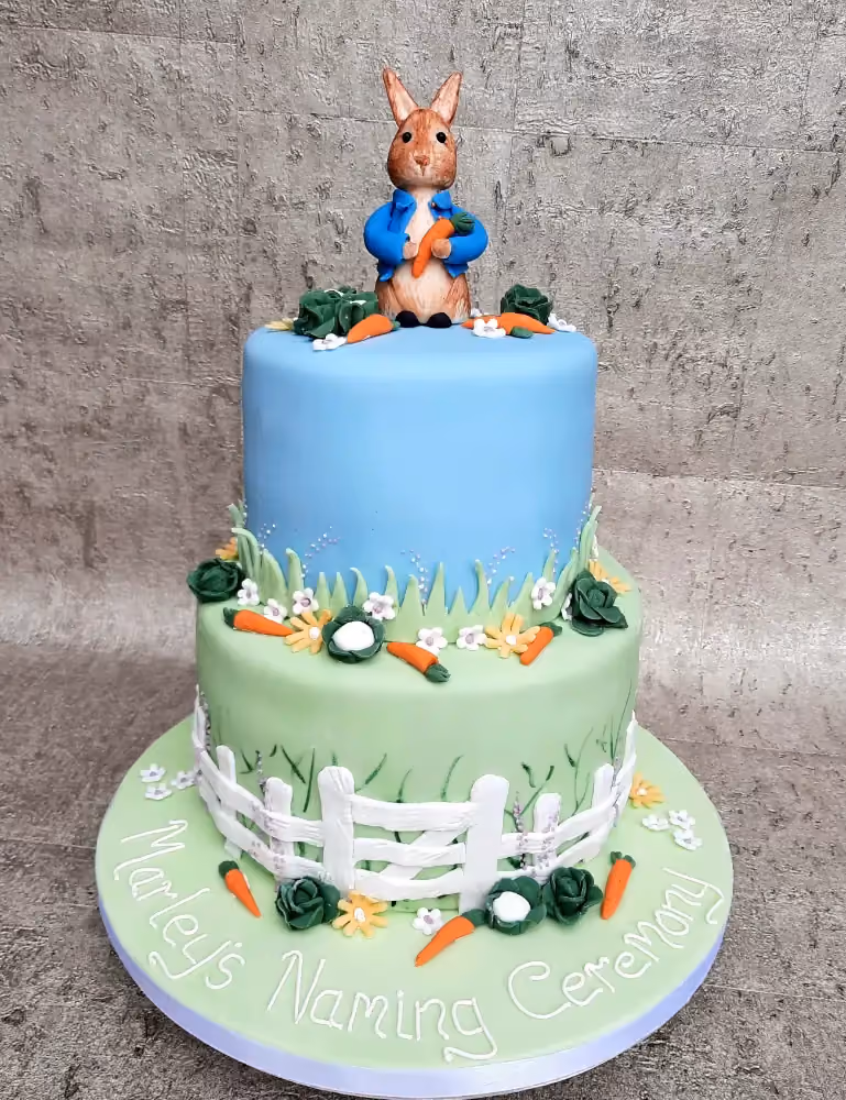 A two-tier Peter Rabbit themed naming ceremony cake, with a blue top tier and a light green bottom tier, decorated with carrots, cabbages, flowers, and a white picket fence, topped with a Peter Rabbit figurine holding a carrot. The cake board is inscribed with "Marley's Naming Ceremony".