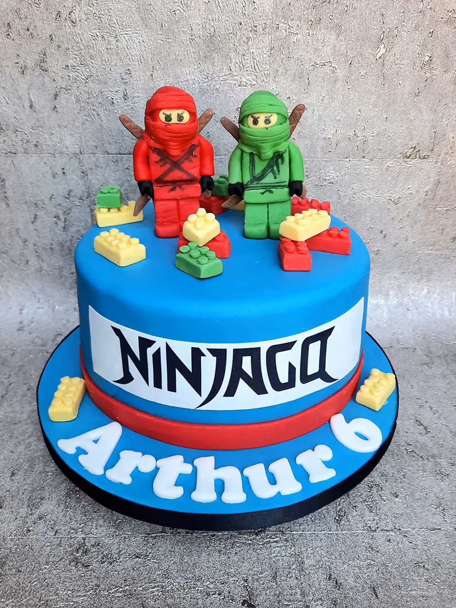 A vibrant blue, single-tier birthday cake decorated with red and green Lego Ninjago characters, surrounded by small edible red, yellow, and green Lego bricks. The side of the cake features a white band with 'NINJAGO' in black lettering, and the blue cake board has 'Arthur 6' written in white fondant letters.