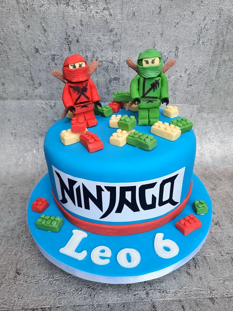 A round, bright blue fondant cake decorated with two fondant Lego Ninjago figures, one red and one green, standing on top. Around the figures are small fondant Lego bricks in red, green, and cream. The side of the cake features a white band with the word 'NINJAGO' in black letters, and on the cake board, in white letters, are the name 'Leo' and the number '6'.