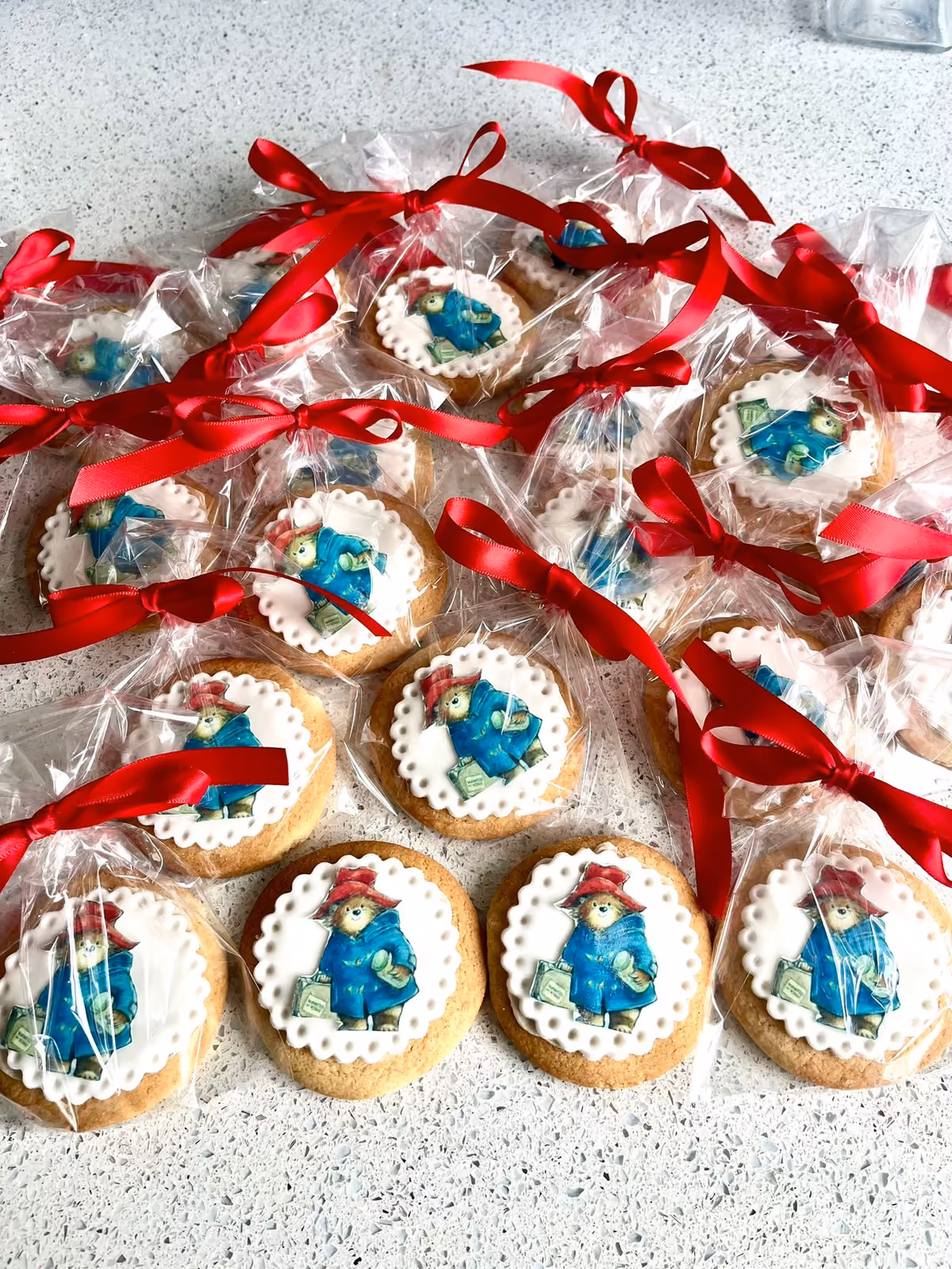 A collection of round cookies, each topped with a white scalloped icing circle featuring an edible image of Paddington Bear, individually wrapped in clear plastic bags tied with red satin ribbons.