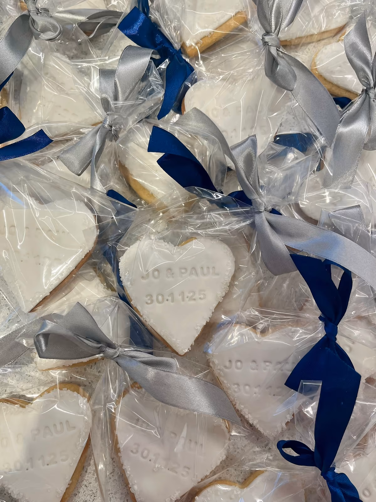 A close-up overhead view of multiple individually wrapped heart-shaped biscuits, each personalized with 'JO & PAUL' and '30.11.25', tied with alternating blue and silver ribbons.