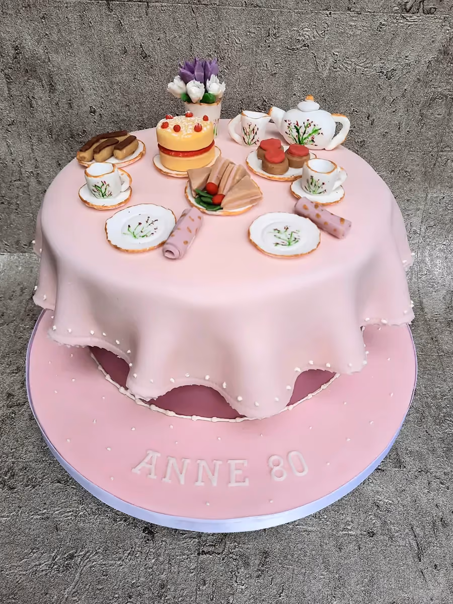 A two-tier pink birthday cake for Anne's 80th, designed as a miniature tea party with a draped tablecloth, tiny cakes, sandwiches, a teapot, cups, and a vase of flowers on top.