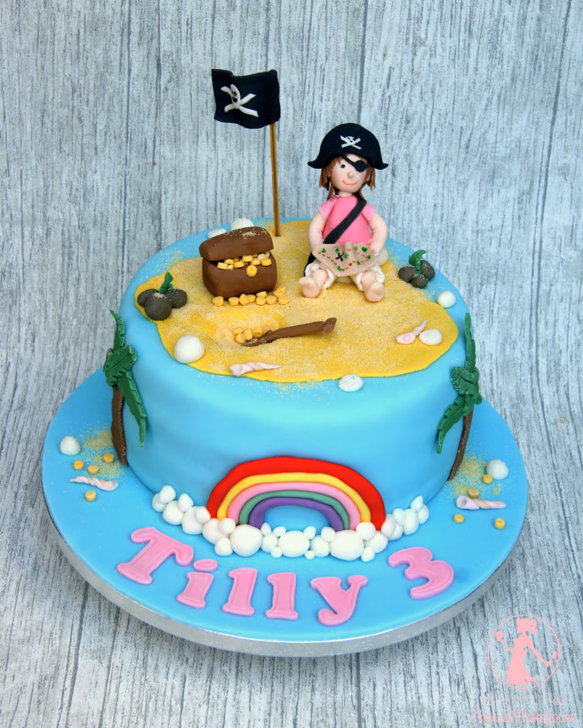 A two-tier pirate island birthday cake featuring a pirate girl, treasure chest, palm trees, and a pirate flag on the top tier, with a rainbow and the name 'Tilly 3' in pink lettering on the bottom tier.
