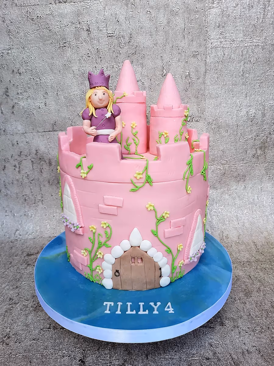 A pink princess castle cake featuring a princess figure on top, complete with turrets, green vines, yellow flowers, and the name 'TILLY4' written on the blue cake board.