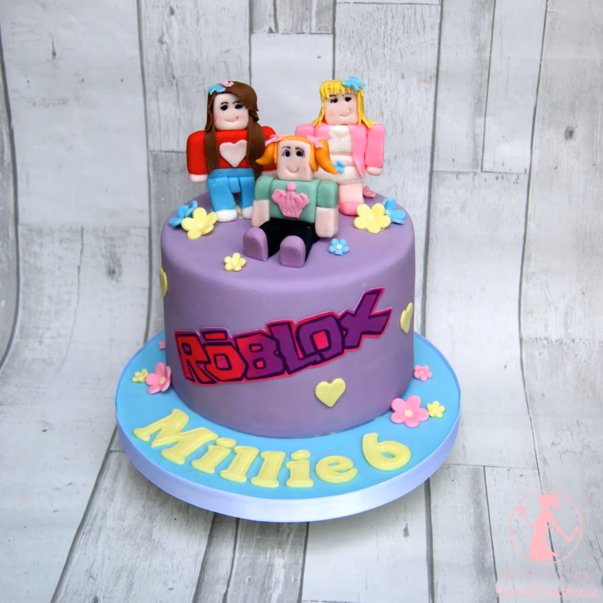 A single-tier purple Roblox birthday cake featuring three fondant Roblox-style girl figures on top, the Roblox logo on the side, and 'Millie 6' in yellow letters on the blue cake board, adorned with small flowers and hearts.