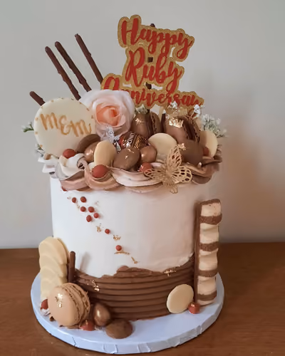 A white and chocolate tiered anniversary cake, adorned with a 'Happy Ruby Anniversary' topper, assorted chocolates, macarons, red berries, a pink artificial rose, and a gold butterfly.